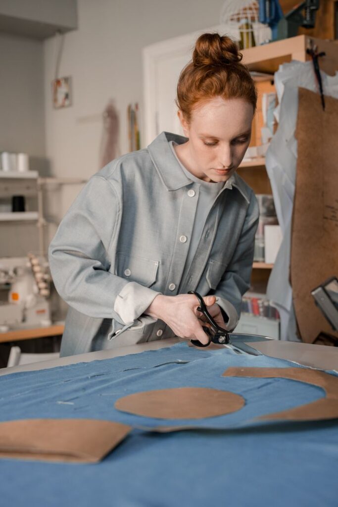 Person in a workshop cutting blue fabric with scissors, surrounded by sewing materials and patterns, focused on crafting or fashion design.