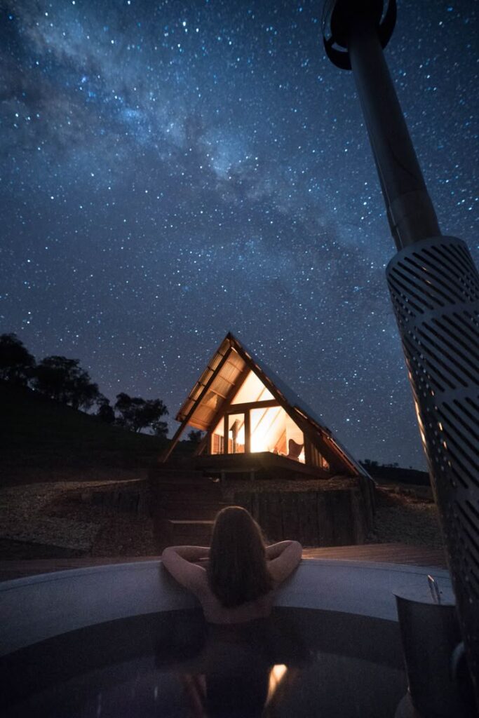 Person relaxing in a hot tub under a starry sky near a cozy, lit A-frame cabin.