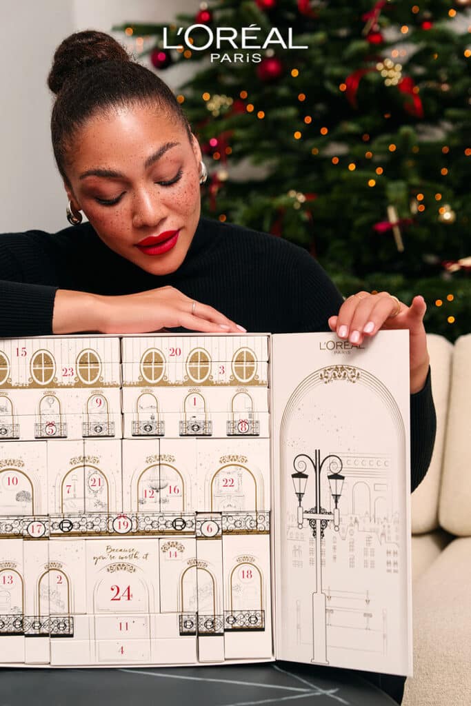 Woman with red lipstick and bun admires L'Oréal Paris advent calendar; festive setting with Christmas tree in the background.