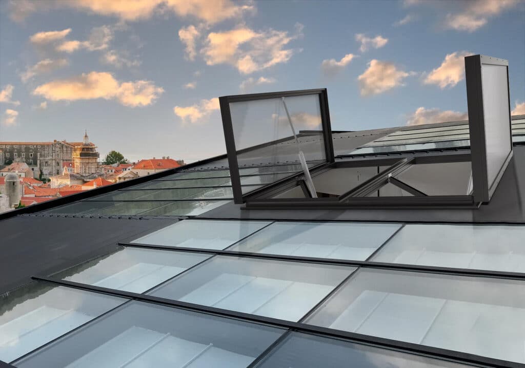 Modern glass rooftop with open skylight, overlooking a historic European cityscape under a vibrant, cloudy sky.