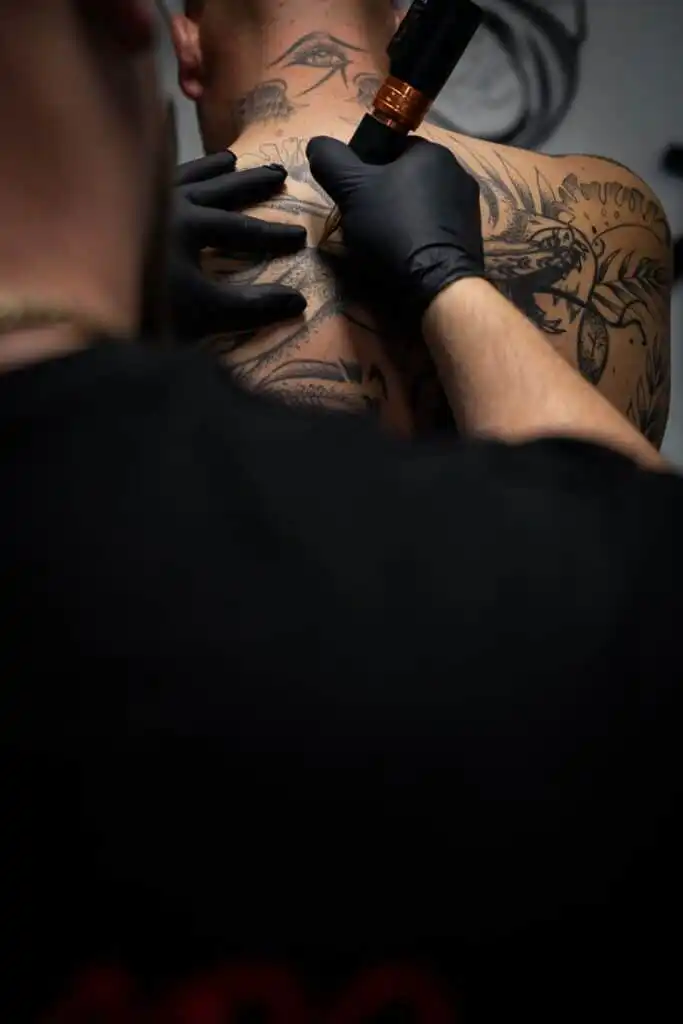 Tattoo artist inks intricate back tattoo using a tattoo machine. Black gloves and detailed tattoo design in progress.