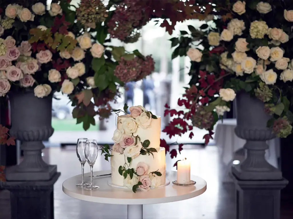 Lancemore-Mansion-Hotel-Werribee-Park-Lost-in-Love-photography__Resampled Elegant wedding cake adorned with white and pink roses, surrounded by lush floral arrangements and accompanied by glasses and a candle. | Sky Rye Design Elegant wedding cake adorned with white and pink roses, surrounded by lush floral arrangements and accompanied by glasses and a candle.