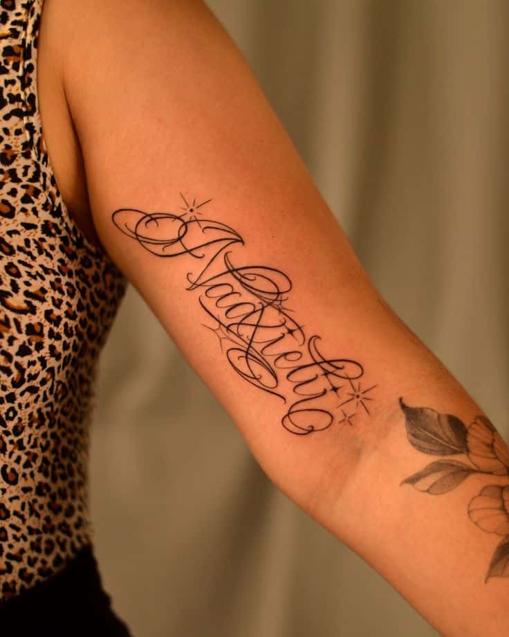 Elegant script tattoo on forearm with floral design, worn by person in leopard-print top, showcasing a blend of personal style and artistry.