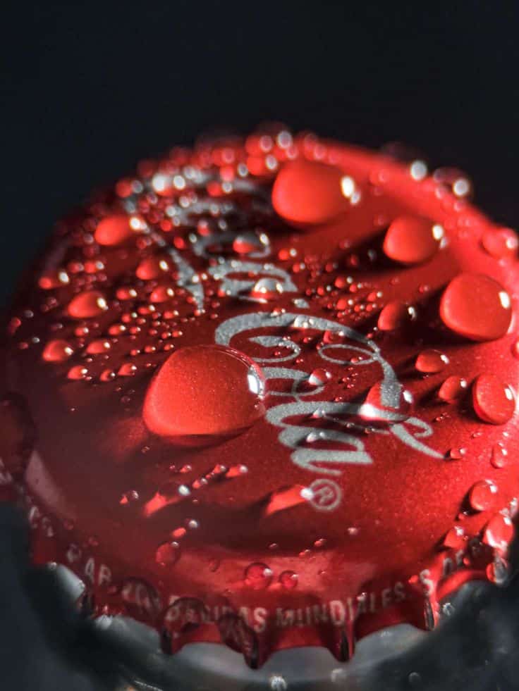 Close-up of a red soda bottle cap with water droplets, highlighting the brand logo, giving a refreshing and chilled appearance.