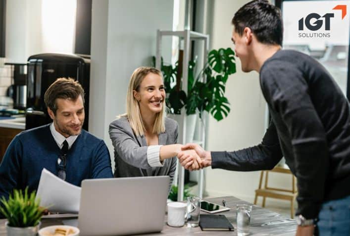 Three colleagues smiling and shaking hands during a meeting in a modern office setting with a laptop and documents on the table.