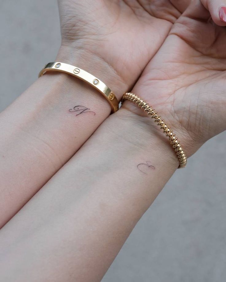 Matching initials tattoo for best friends_ Two wrists with delicate letter tattoos, each adorned with a gold bracelet; minimalistic and elegant wrist art. | Sky Rye Design Two wrists with delicate letter tattoos, each adorned with a gold bracelet; minimalistic and elegant wrist art.