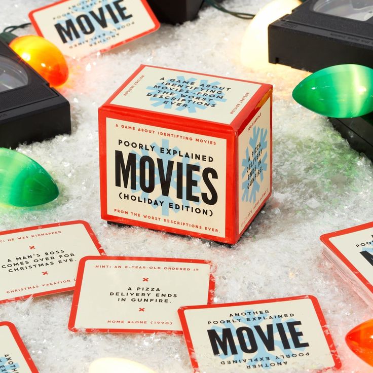 Board game Poorly Explained Movies (Holiday Edition) with movie cards and VHS tapes on snowy surface, surrounded by lights.