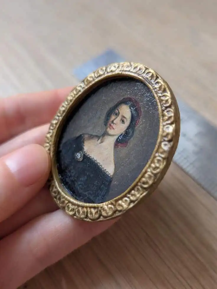 Close-up of a hand holding a small, round antique portrait of a woman in a gilded frame, resting on a wooden surface.