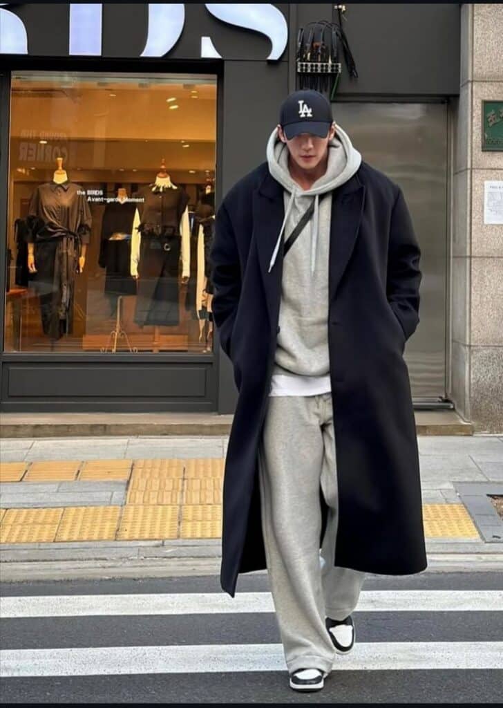 Man in a black coat and gray outfit, baseball cap, walking across a street with a fashion store in the background. Urban street style.