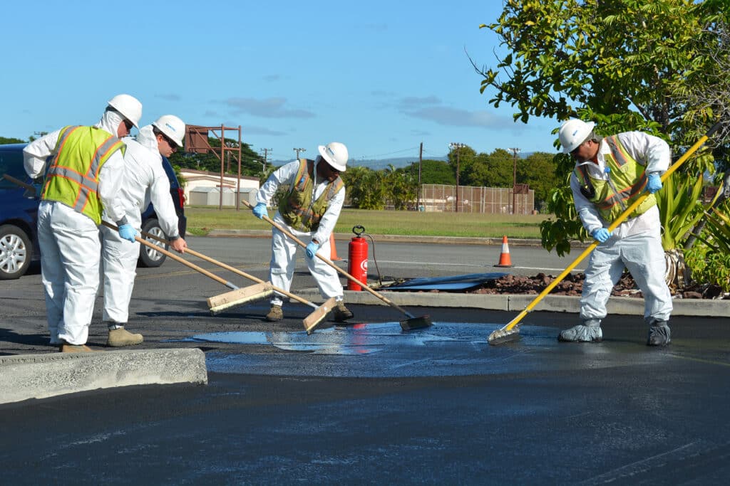 Construction workers in safety gear paving a road with brushes and tools under a clear blue sky.