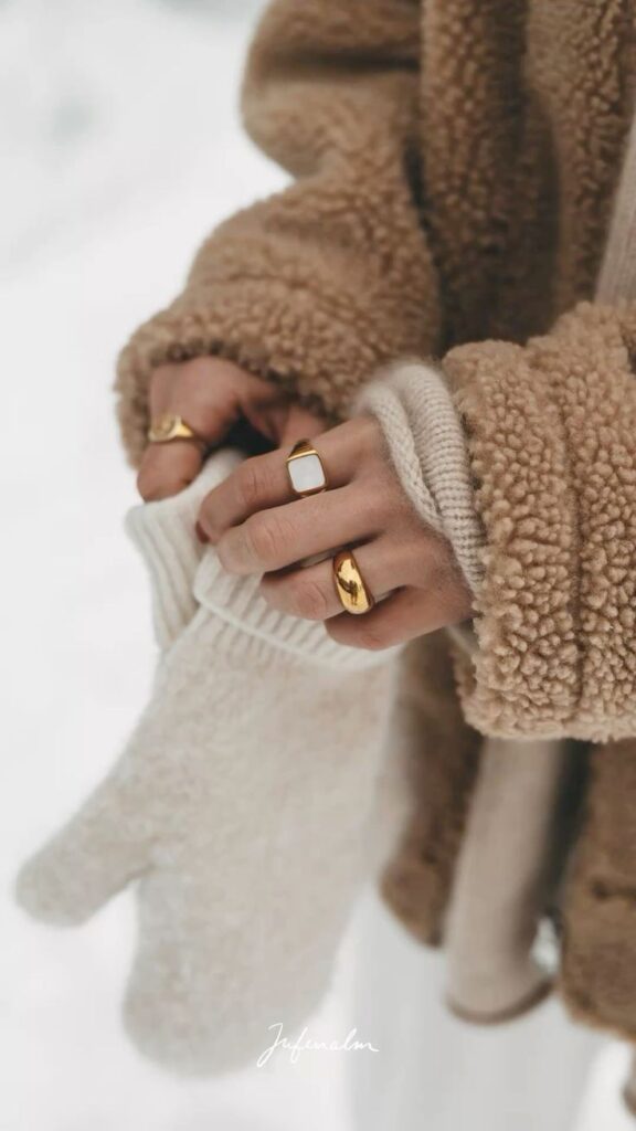 NOOR Cozy winter fashion, person in a sherpa jacket holding knit gloves, wearing gold rings. Warm, stylish layering for cold weather. | Sky Rye Design Cozy winter fashion, person in a sherpa jacket holding knit gloves, wearing gold rings. Warm, stylish layering for cold weather.