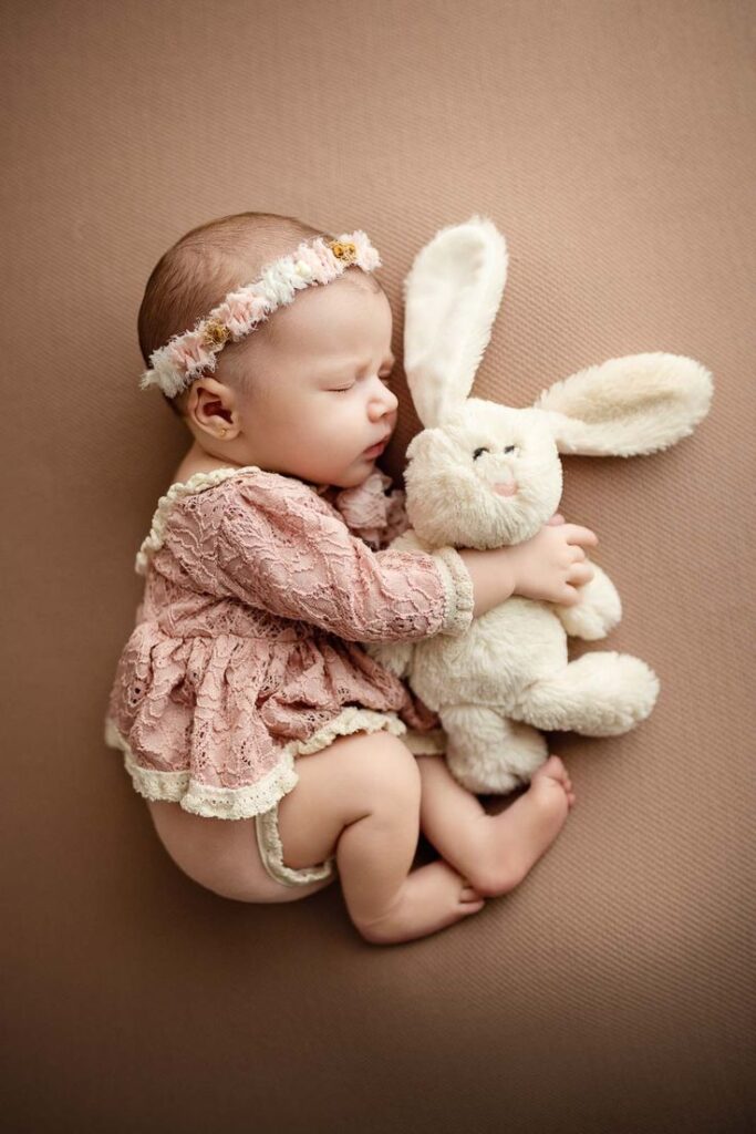 Evoto Sleeping baby in pink lace dress cuddles a plush bunny on soft brown backdrop, wearing a floral headband. Cozy and serene moment. | Sky Rye Design Sleeping baby in pink lace dress cuddles a plush bunny on soft brown backdrop, wearing a floral headband. Cozy and serene moment.