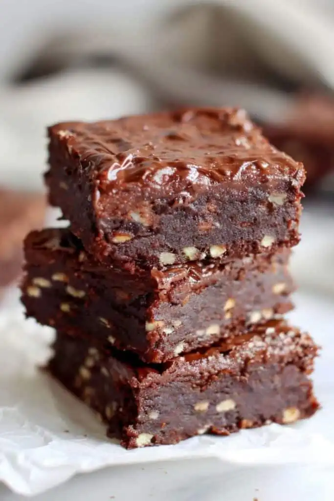 Stack of three rich, fudgy brownies with nuts on parchment paper, highlighting their glossy surface and gooey texture.