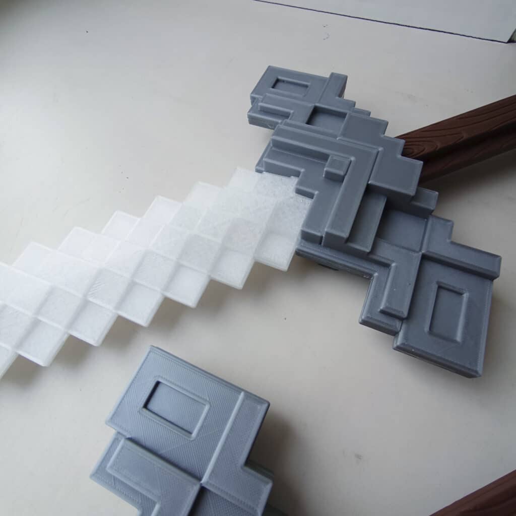 O7kECyPOwq4 3D-printed pixelated sword in gray and white, showcasing a retro, blocky design on a light background. Ideal for gaming fans. | Sky Rye Design 3D-printed pixelated sword in gray and white, showcasing a retro, blocky design on a light background. Ideal for gaming fans.