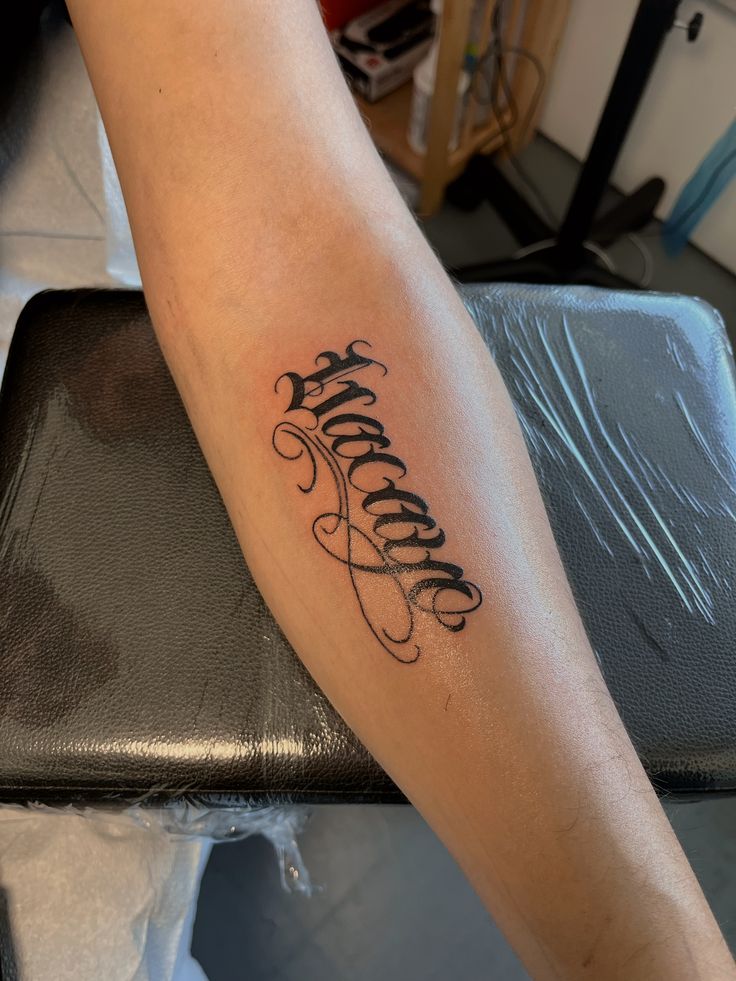 Forearm tattoo with intricate lettering design, showcasing artistic black ink on skin, resting on a black cushioned surface in a tattoo studio.
