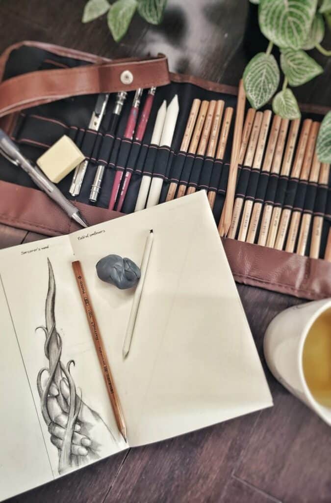 Artist's sketchbook with pencils, eraser, and a pencil case on a wooden table, featuring a detailed drawing and green plants nearby.