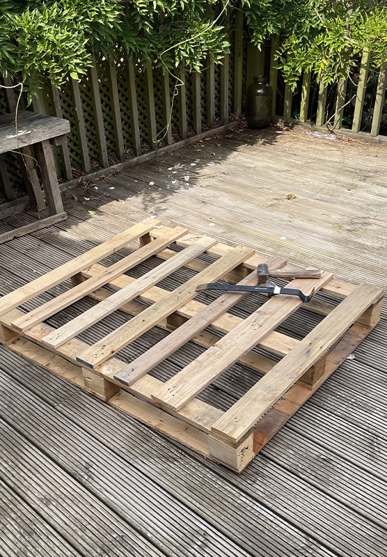 Pallet-before Wooden pallet with tools on a deck, surrounded by greenery. Ideal for DIY projects or upcycling ideas in a garden setting. | Sky Rye Design Wooden pallet with tools on a deck, surrounded by greenery. Ideal for DIY projects or upcycling ideas in a garden setting.