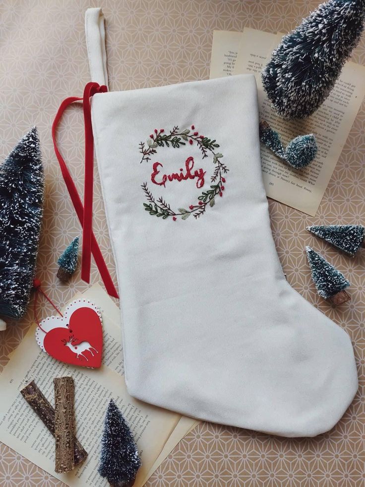 Personalized Family Christmas Santa stocking Custom name Stocking Linen hand embroidered stocking Kids Christmas stocking White Christmas stocking with personalized embroidery, surrounded by mini trees, books, and heart ornament, on a patterned background. | Sky Rye Design White Christmas stocking with personalized embroidery, surrounded by mini trees, books, and heart ornament, on a patterned background.