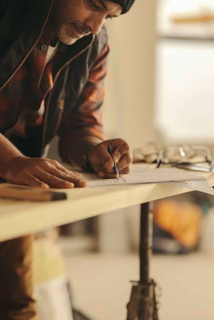 Planning a Home Renovation_ Read This First __ Man focused on drawing on a large sheet of paper at a workbench, wearing a beanie and plaid shirt, with measuring tools nearby. | Sky Rye Design Man focused on drawing on a large sheet of paper at a workbench, wearing a beanie and plaid shirt, with measuring tools nearby.