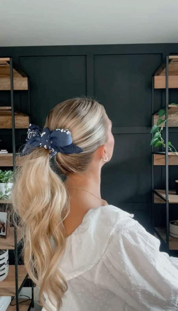 Ponytail Bow 🎀 Woman with a wavy blonde ponytail tied with a navy bow, wearing a white blouse, stands in front of dark shelving with plants. | Sky Rye Design Woman with a wavy blonde ponytail tied with a navy bow, wearing a white blouse, stands in front of dark shelving with plants.