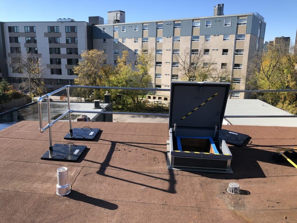 Rooftop with safety rails and open hatch, overlooking city buildings and trees on a clear, sunny day.