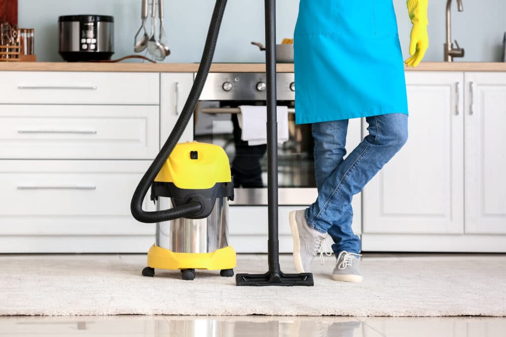 Person in apron stands with vacuum cleaner in a bright kitchen.