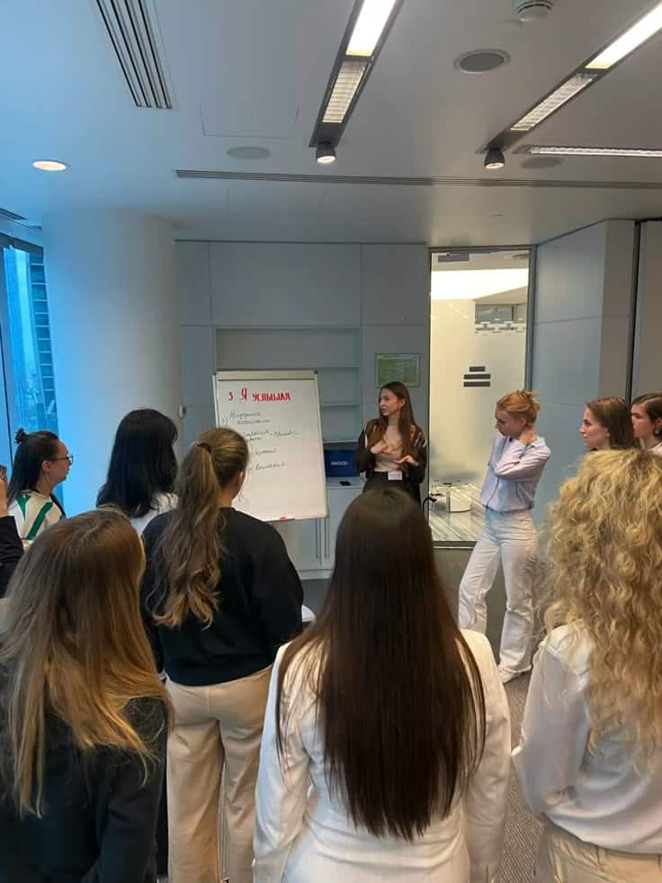 A group of people in an office setting attend a presentation with a speaker next to a flip chart, engaged in a discussion.