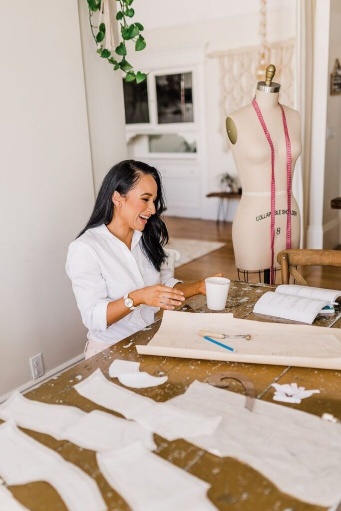 Rising Star_ Jessica Silvas Fashion Designer Journey - Brand Photoshoot New Business Launch Designer smiling at her workstation with a dress form, sewing patterns, and a cup of coffee, enjoying a creative break. | Sky Rye Design Designer smiling at her workstation with a dress form, sewing patterns, and a cup of coffee, enjoying a creative break.