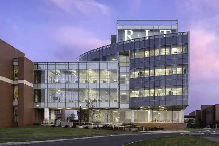 Modern glass building with RIT sign at sunset, featuring illuminated interior and a sleek architectural design against a purple sky.