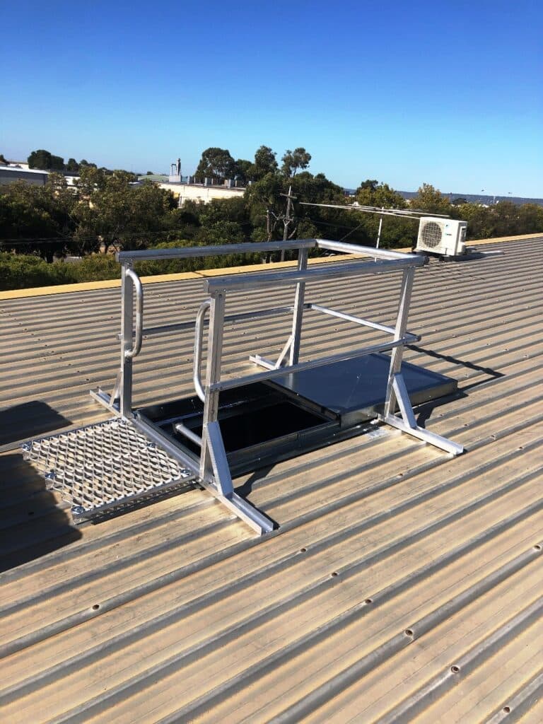 Rooftop safety hatch with railing on corrugated metal, providing secure roof access. Blue sky and green trees in the background.
