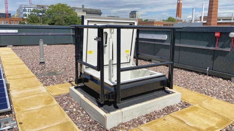 Rooftop access hatch with safety railing, surrounded by gravel and solar panels, providing secure entry to roof area under blue sky.
