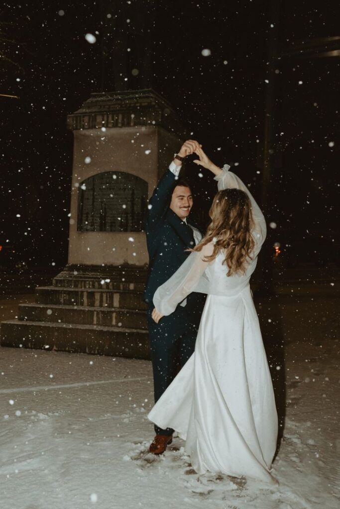 Sabina + Chase Dance in the Snow _ Spokane Club Wedding Bride and groom dancing joyfully in falling snow at night, surrounded by soft lights and a romantic atmosphere. | Sky Rye Design Bride and groom dancing joyfully in falling snow at night, surrounded by soft lights and a romantic atmosphere.
