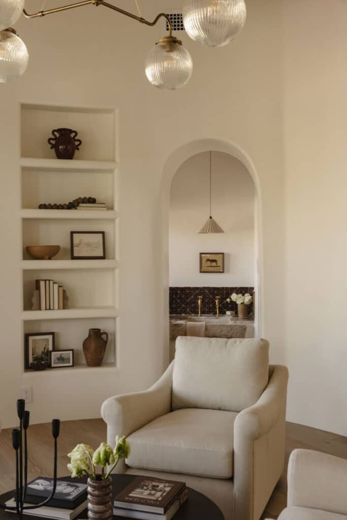 Santa Fe Hacienda _ Living Room Cozy living room with beige armchair, bookshelves, elegant lighting, and modern decor, featuring a view into a stylish, earthy-toned room. | Sky Rye Design Cozy living room with beige armchair, bookshelves, elegant lighting, and modern decor, featuring a view into a stylish, earthy-toned room.