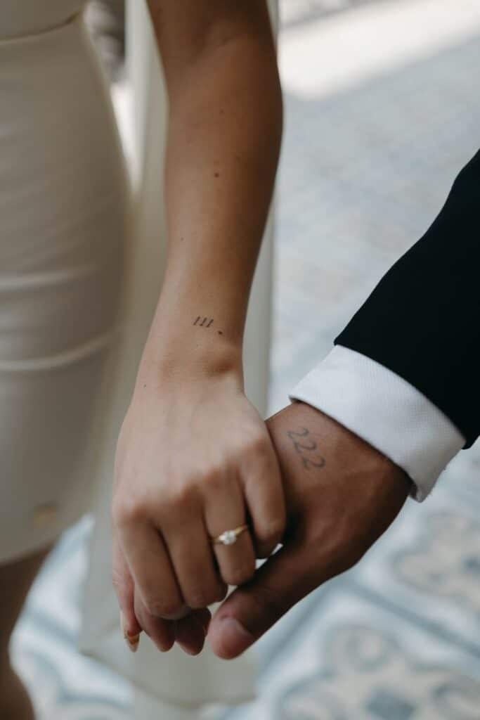 Couple holding hands with matching tattoos, woman in white dress, man in suit. Tender moment symbolizing unity and love.