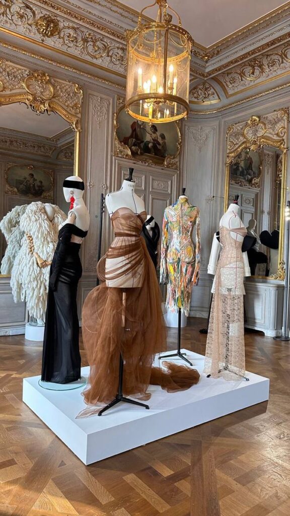 Schiaparelli Elegant room with mannequins showcasing four designer dresses, under a grand chandelier and ornate decor. Perfect for fashion inspiration. | Sky Rye Design Elegant room with mannequins showcasing four designer dresses, under a grand chandelier and ornate decor. Perfect for fashion inspiration.