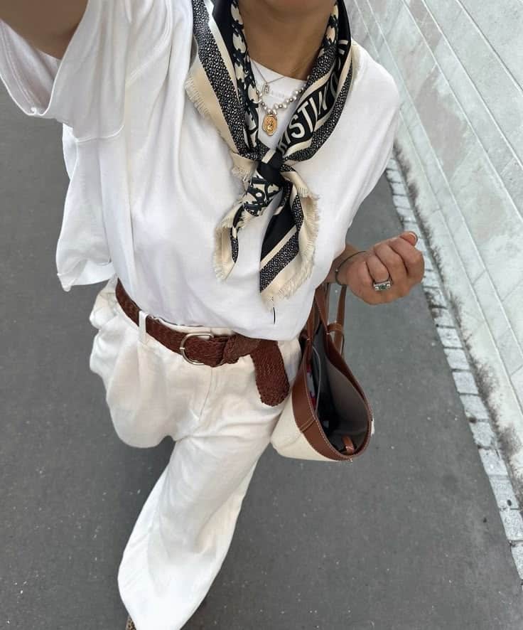 Stylish outfit with white tee, patterned scarf, brown belt, and wide-leg pants, accessorized with layered necklaces and a leather tote.