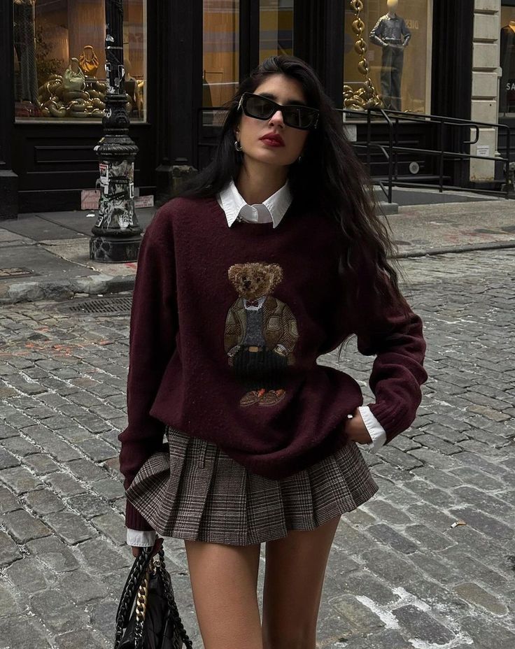 Skirt outfit __Aesthetic outfit __Casual outfit __Long sleeves outfit __White shirt outfit __Button up shirt outfit __Black purse __Sweatshirt outfit __Black glasses __Short skirt outfit Fashionable woman wearing a bear-graphic sweater, plaid skirt, and sunglasses on a city street, embodying chic autumn style. | Sky Rye Design Fashionable woman wearing a bear-graphic sweater, plaid skirt, and sunglasses on a city street, embodying chic autumn style.
