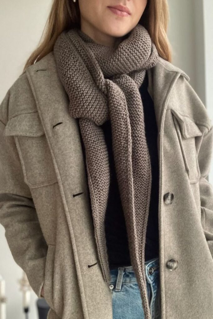 Woman in a stylish beige coat and brown knitted scarf, wearing blue jeans, showcases a chic fall fashion outfit.