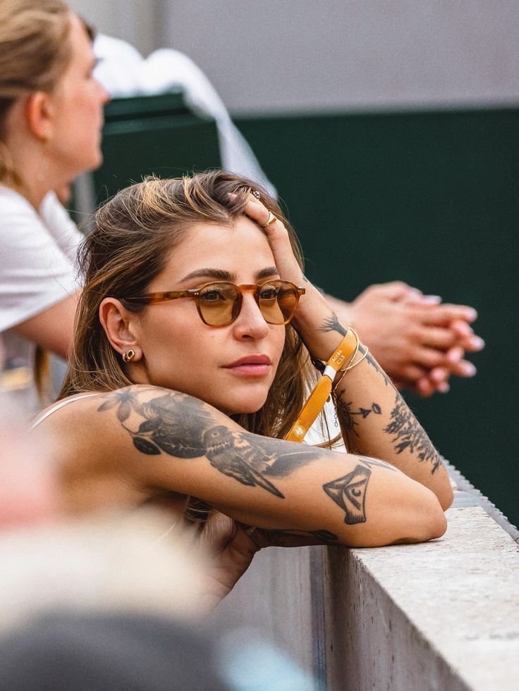 Woman with tattoos and sunglasses leans on a barrier, gazing thoughtfully. Relaxed outdoor scene with a focus on her artistic ink.