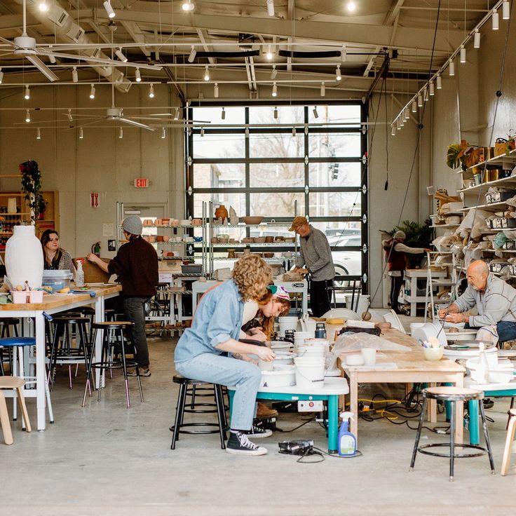 People engaged in pottery making inside a well-lit studio, with shelves of ceramics and large windows in the background.
