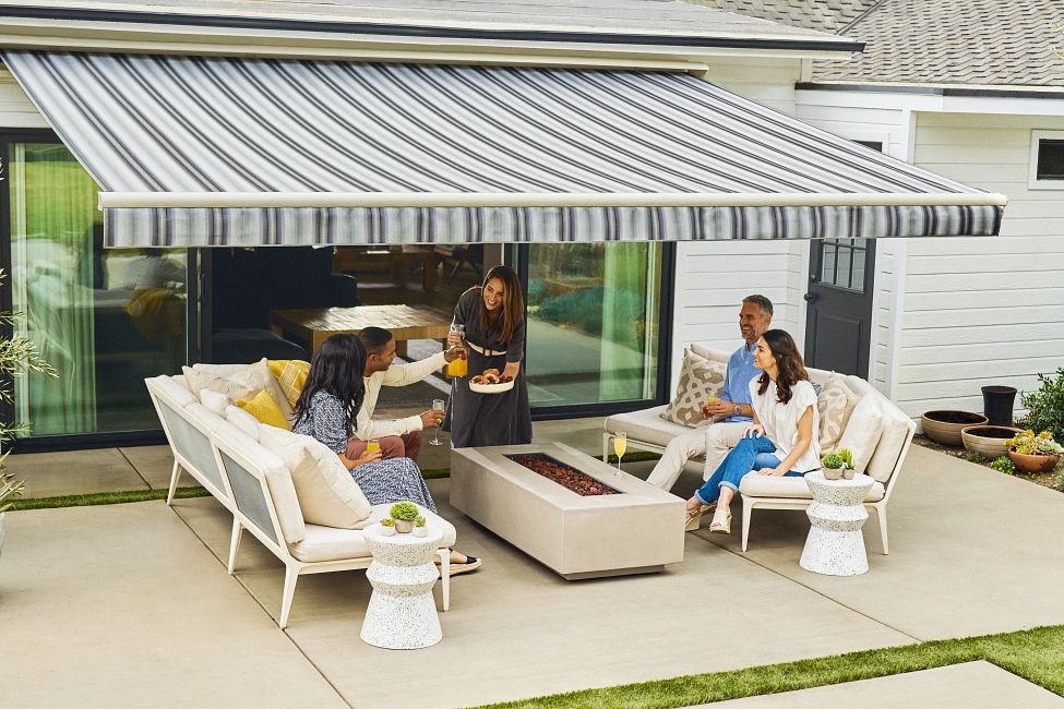 SunSetter-LS21-PlatMotorPRO-StormStripe-17 Group of people enjoying a sunny outdoor gathering under a striped retractable awning, with drinks and snacks on a patio lounge set. | Sky Rye Design Group of people enjoying a sunny outdoor gathering under a striped retractable awning, with drinks and snacks on a patio lounge set.