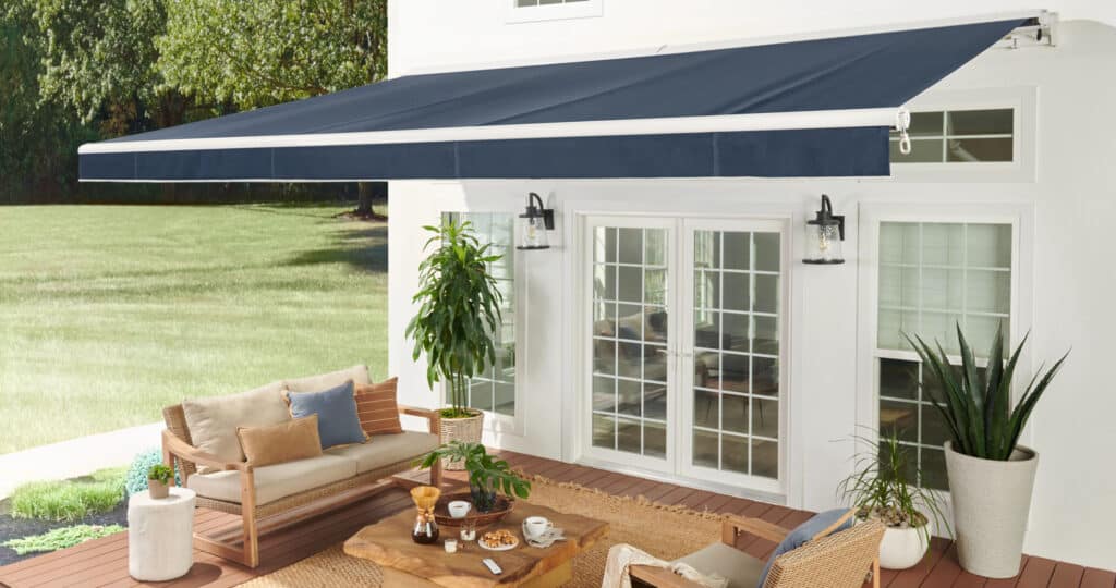 SunSetter-PA22-Model-Fabric-for-the-Sunsetter-V3-3000x1582 Outdoor patio with blue retractable awning, modern furniture, plants, and a wooden table set against a backdrop of green lawn and trees. | Sky Rye Design Outdoor patio with blue retractable awning, modern furniture, plants, and a wooden table set against a backdrop of green lawn and trees.