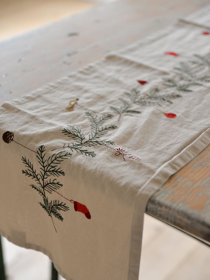 TABLE RUNNER SPRUCE WREATH Festive embroidered table runner with pine branch design and red accents, perfect for holiday dining decor. | Sky Rye Design Festive embroidered table runner with pine branch design and red accents, perfect for holiday dining decor.