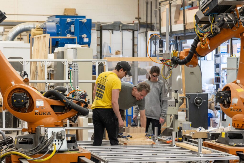TCAUP_FABLab_20240415-37-scaled People collaborating with industrial robots in a manufacturing workshop, surrounded by machinery and equipment. | Sky Rye Design People collaborating with industrial robots in a manufacturing workshop, surrounded by machinery and equipment.