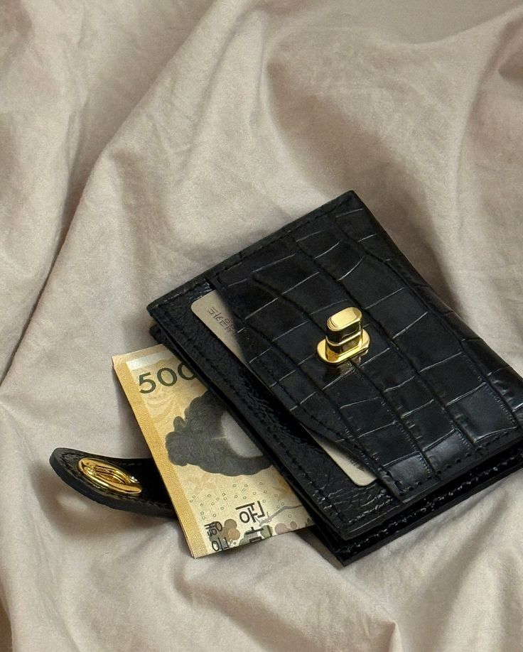 Black leather wallet with gold clasp, featuring a 500 currency note, stylishly placed on beige fabric background.