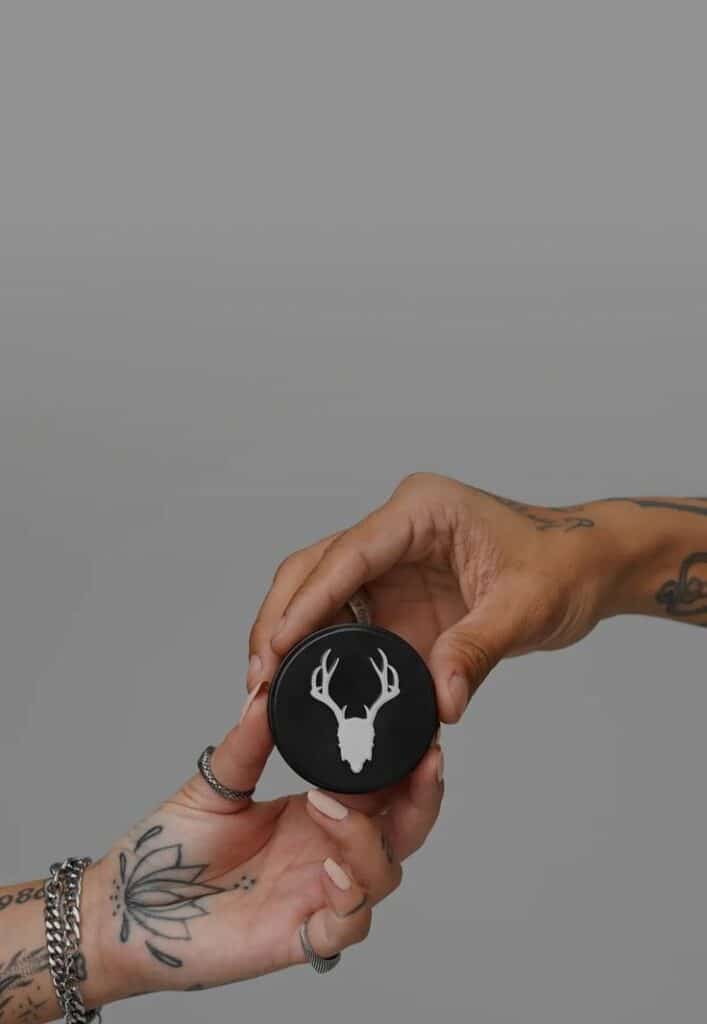 Tattoo Care Products _ All Your Aftercare Needs Hands holding a black round container with a white deer logo, against a gray background, showcasing tattoos and jewelry. | Sky Rye Design Hands holding a black round container with a white deer logo, against a gray background, showcasing tattoos and jewelry.