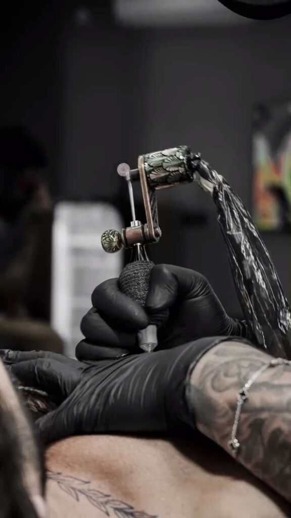 Tattoo machine Tattoo artist wearing black gloves, using a tattoo machine to ink a detailed design on a person's skin. | Sky Rye Design Tattoo artist wearing black gloves, using a tattoo machine to ink a detailed design on a person's skin.
