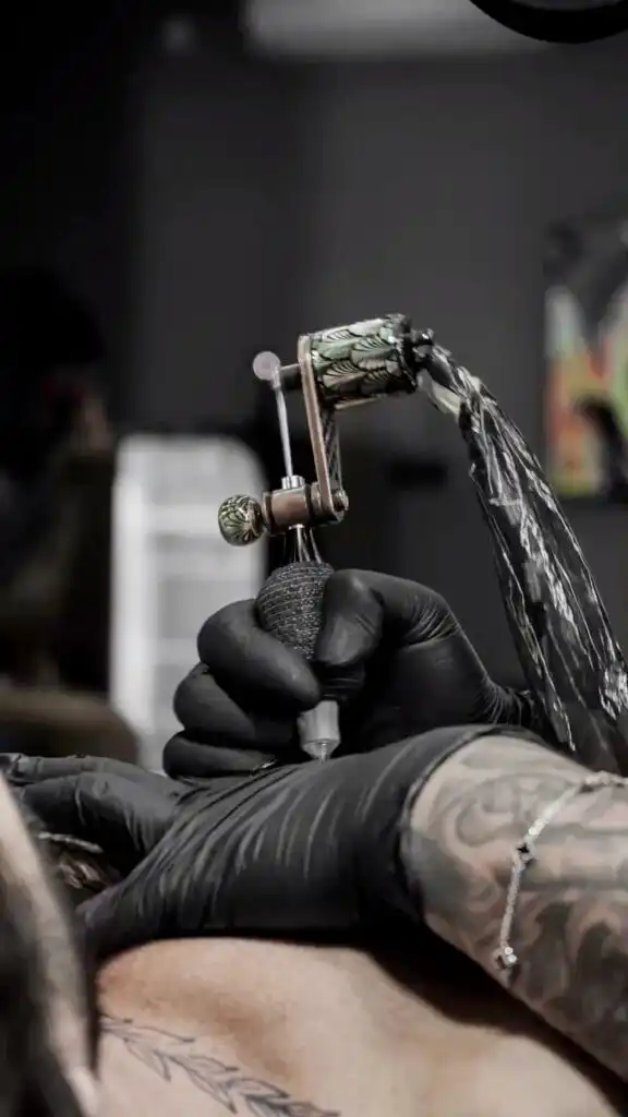 Tattoo artist wearing black gloves, using a tattoo machine to ink a detailed design on a person's skin.