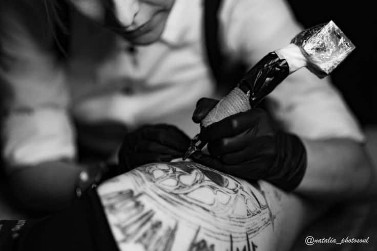 Tattoo studio Tattoo artist skillfully inking a detailed design on a client's leg in a studio setting. Black and white close-up image. | Sky Rye Design Tattoo artist skillfully inking a detailed design on a client's leg in a studio setting. Black and white close-up image. how much is a tattoo gun