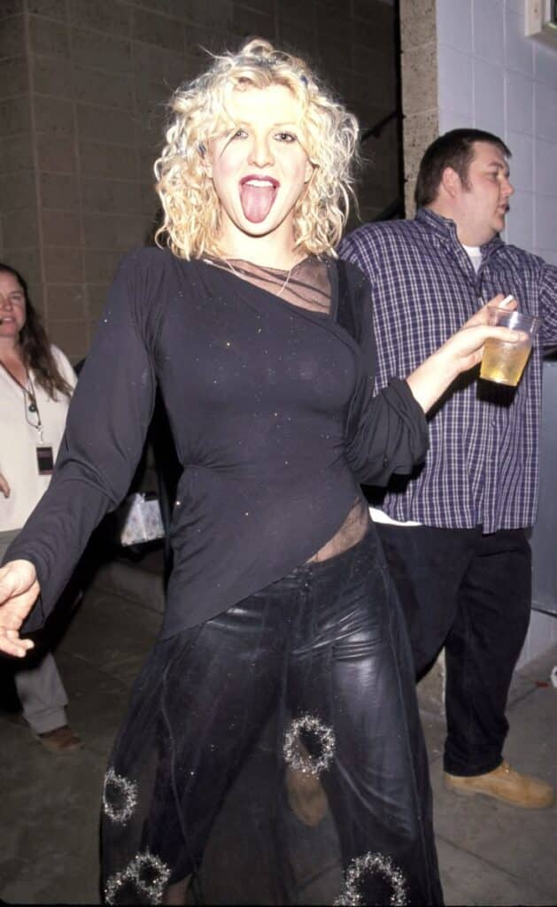 Woman in black outfit playfully sticks out tongue while holding drink, surrounded by people at an event. Fun, lively atmosphere.